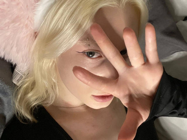 Close-up photo of Enigma-hot with platinum blonde hair, covering one eye with a hand. Pale skin, visible eye makeup, black top, light pink furry hat, intense gaze, sexual tension.