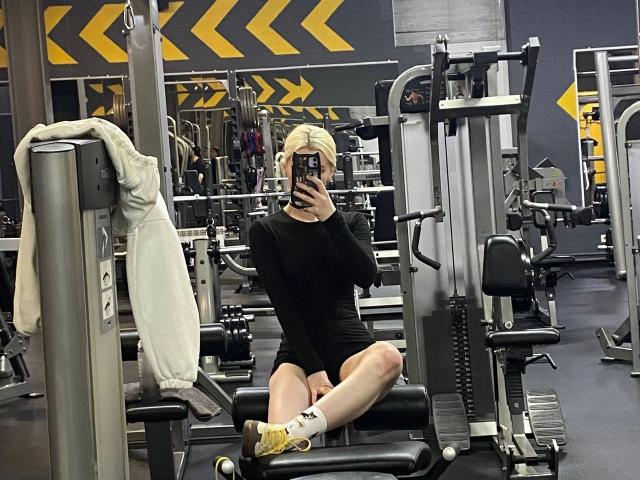 Een vrouw met blond haar, die er aantrekkelijk uitziet, zit op een fitnessapparaat, haar benen gespreid, en draagt zwarte shorts en gele sokken. Ze houdt een telefoon vast. Spiegelfoto, gespierd postuur, sportschoolomgeving, gele veiligheidsstrepen op de achtergrond..