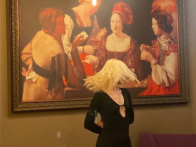 A painting of Enigma-hot figures in 17th-century attire, intense expressions, red and gold colors. A real-life Enigma-hot with blonde hair and black dress in front, head turned away. Sexual tension.