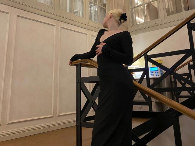 Enigma-hot stands near a staircase, leaning on a wooden railing with a black dress, long sleeves, and a black hair bow. Fucking sexy, black outfit accentuates her curvy figure.