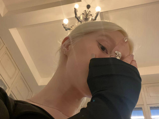 Close-up selfie of Enigma-hot, a blonde woman with fair skin, covering her mouth with a black-gloved hand. She has large, black-rimmed glasses, and a chandelier is in the background. Fucking intense gaze.