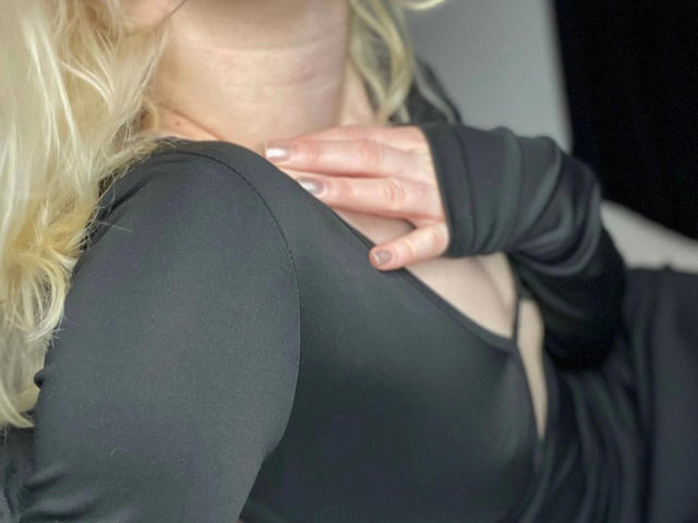 Primo piano dei capelli biondi e della maglia nera di Enigma-hot, una mano che stringe il petto, mostrando il décolleté, la pelle esposta, un leggero rossore sulle guance. collo, luce soffusa, ombre, intimo, sensuale, allusivo, tensione erotica.