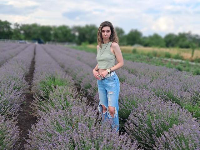 Sanella se tient dans un champ de lavande, vêtue d'un débardeur vert clair et de jeans bleus déchirés. Elle a des cheveux bruns, ondulés et humides, et un regard confiant et séduisant. De la lavande sauvage l'entoure..