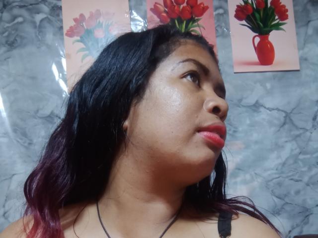 MariahSquirting69 tiene el cabello oscuro y húmedo, piel brillante y lápiz labial rojo, y mira hacia arriba con una ligera sonrisa. El fondo tiene una textura de mármol y dos jarrones con flores rojas en la pared..