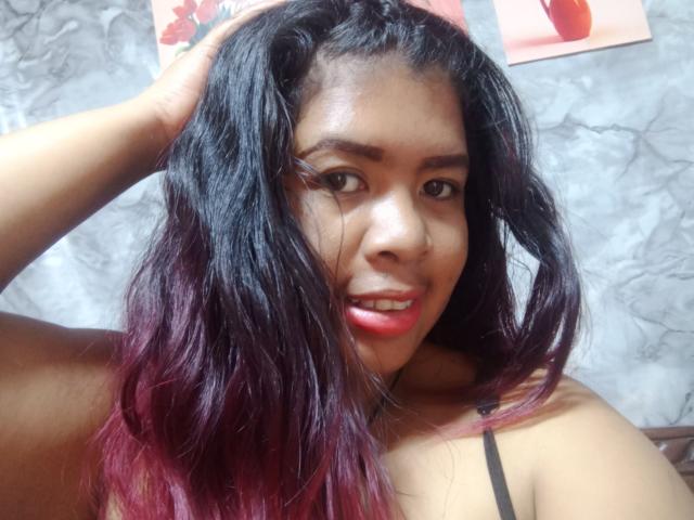 Close-up of MariahSquirting69, a curvy woman with dark, wavy hair and red tips, wearing a strap top, hand in hair, lips slightly parted, red lipstick, bright eyes, light-skinned, sexual tension.