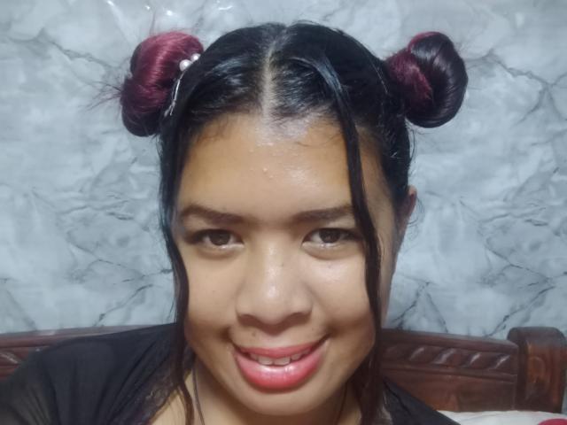 Close-up of MariahSquirting69, an Asian woman with shiny skin, black hair in two maroon buns, and red lipstick, smiling directly at the camera with visible sexual tension in her gaze.