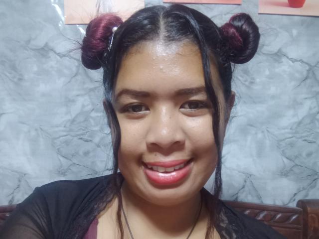 Close-up of MariahSquirting69, an Asian woman with shiny skin, black hair in double buns, red lipstick, smiling, wearing a black top. Marble background, soft lighting, slight gloss on face.