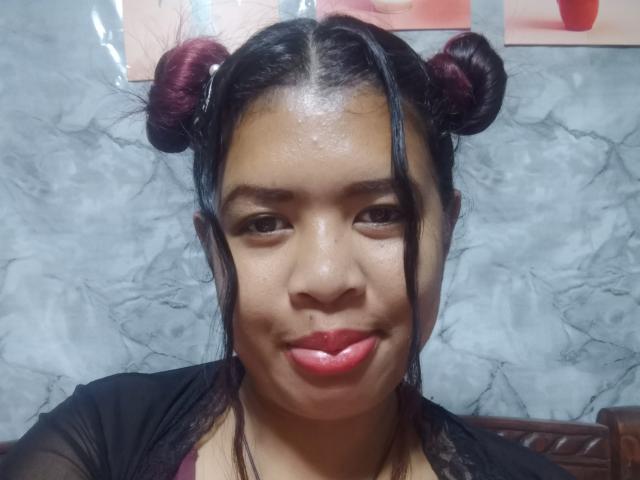 Close-up of MariahSquirting69, an Asian woman with glossy skin, dark hair in two buns, red lipstick, and a black top. Shiny face, sexy expression, background with marble texture.