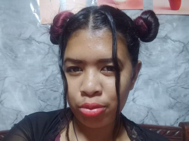 Close-up of MariahSquirting69 with dark hair in two maroon buns, glossy skin, red lipstick, black top, marble wall background. Shiny face, serious expression, slight sexual tension, head slightly tilted.