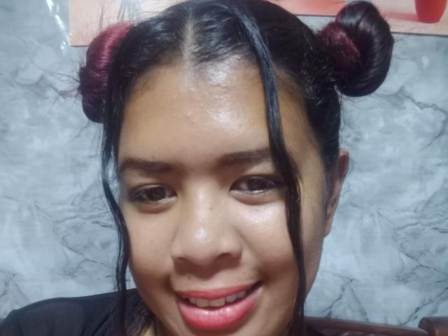 Close-up of MariahSquirting69, Asian woman with shiny skin, black hair in double buns, red lipstick, slight smile, glossy eyes, marble background. Her expression conveys subtle sexual tension.