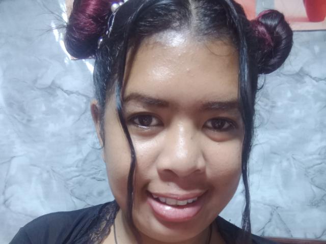 Close-up of MariahSquirting69, an Asian woman with shiny skin, black hair in two red-dyed buns, smiling slightly, visible wetness on her face, wearing a black shirt, against a textured white background.