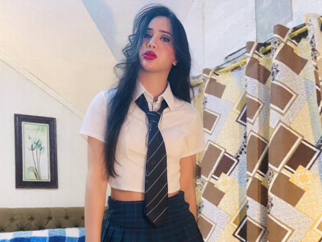 DominantGoddess mit langen, schwarzen Haaren, rotem Lippenstift, einem weißen Crop-Top, einer schwarzen Krawatte und einem karierten Rock. Sie steht in einem hell erleuchteten Raum mit einem gemusterten Vorhang. Verdammt heiß..