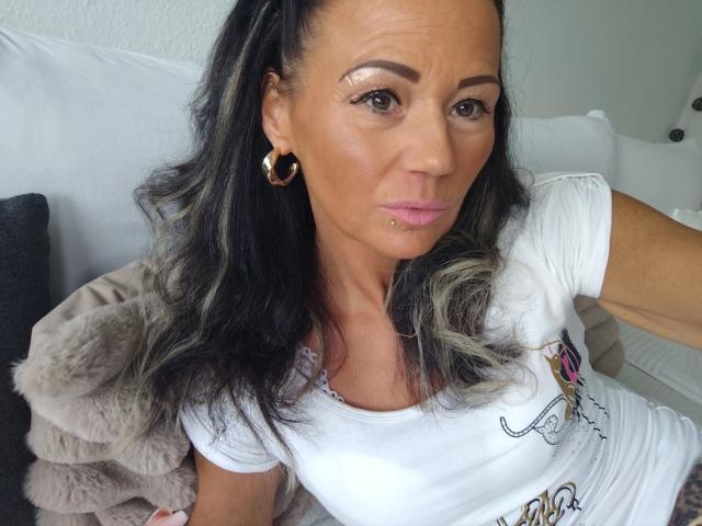 Close-up foto van Miss-Lara, een aantrekkelijke vrouw met donker haar en een piercing in haar onderlip, gekleed in een witte blouse en een gouden oorring. oorringen, zittend op een witte bank met een grijze wollen deken..