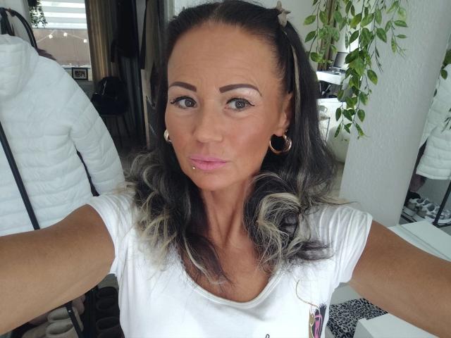 Photographie d'une femme d'âge moyen, Miss-Lara-hot, avec de longs cheveux bruns ondulés, portant un t-shirt blanc et des boucles d'oreilles cercles. et un piercing au lobe, elle tend les bras pour un selfie, ce qui révèle une tension sexuelle..