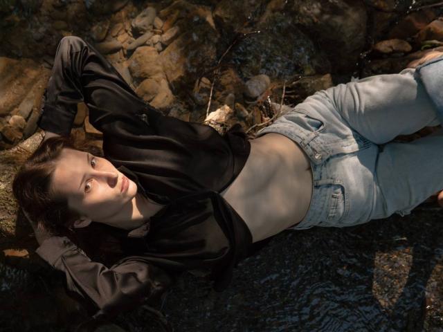 Priscila Vallas est allongée sur un sol rocheux, vêtue d'un haut noir à décolleté plongeant et d'un jean gris clair. Ses bras sont derrière sa tête, et les ombres mettent en valeur sa silhouette. La lumière du soleil sur sa peau.