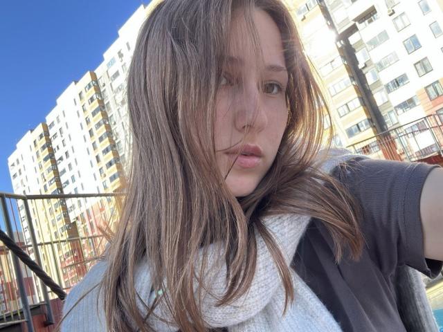 Priscila Vallas ha lunghi capelli castani, leggermente spettinati, che coprono parte del suo viso. Indossa una sciarpa bianca e una maglia nera, ed è in piedi di fronte a degli edifici residenziali illuminati dal sole. Uno sguardo incredibilmente intenso..