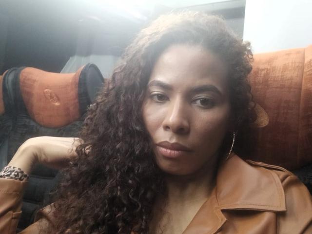 Rebeca Duval, di carnagione scura con capelli neri ricci, indossa una giacca di pelle marrone, seduta su un sedile dell'auto con un cuscino arancione. inquadratura, sguardo diretto verso la telecamera, espressione neutra, luce naturale soffusa.