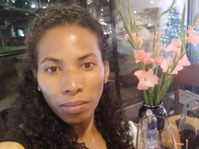 Fotografia de Rebeca Duval, com cabelos cacheados escuros, olhos castanhos e expressão neutra, sentada à mesa com flores rosa. vaso de vidro, copo de vinho, luz fraca, fundo com luzes desfocadas. Tensão sexual.