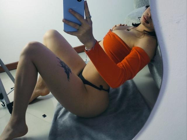 NiickyySweet reclines on a gray blanket, wearing an orange off-shoulder top and black thong, holding a phone. Tattoo on left thigh. Legs crossed, head tilted back. Focused selfie.