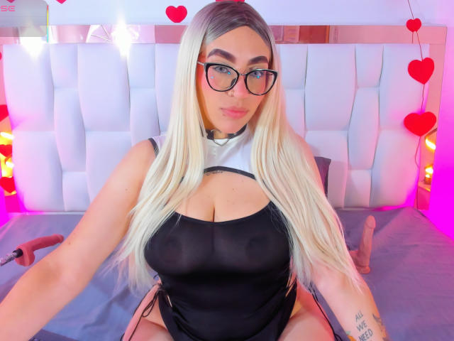 CamilleJuicyHot, formosa, indossa un abito nero con aperture, occhiali, capelli biondi, seduta sul letto, con una testiera bianca imbottita, di colore rosso. cuori, sex toy sul letto, tatuaggio sull'avambraccio sinistro, sguardo seducente, luci al neon brillanti.