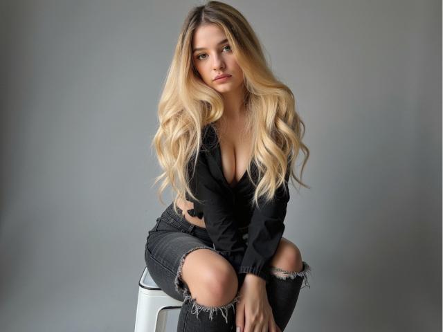 EvelineAngel, avec de longs cheveux blonds, assise sur un tabouret blanc, portant un haut noir à décolleté et un jean noir déchiré. Elle regarde l'appareil photo avec une expression sexy et confiante. Fond gris.
