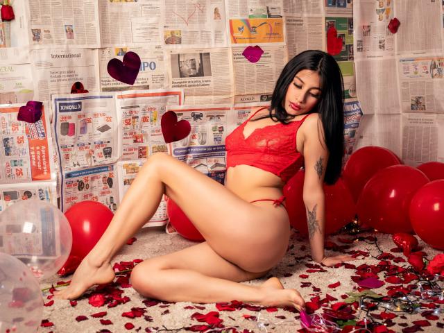 Madhison, con cabello negro y tatuajes, está sentada en el suelo, cubierto de pétalos de rosa roja y globos, vestida con encaje rojo. ropa interior femenina. Sus piernas están separadas, los ojos cerrados, irradiando tensión sexual..