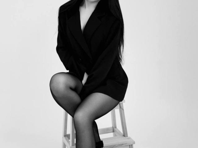 Black and white photograph of AnnabellDeltoro sitting on a wooden stool, legs crossed, wearing a tight black blazer and short skirt, emphasizing her slim figure, with long dark hair.