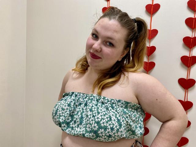 Ailia, a curvy woman with fair skin and brown hair in a ponytail, wears a green and white floral strapless top. Red heart decorations hang in the background. She smiles slightly, exuding casual sexual allure.