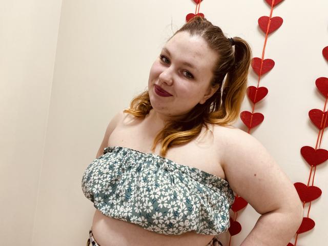 Ailia, a curvy woman with fair skin and brown hair in a ponytail, wears a floral strapless top, standing against a wall with red heart decorations. Her expression is seductive.