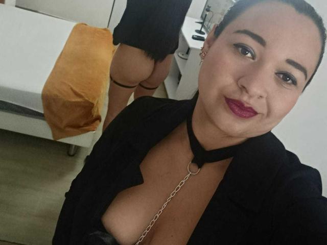 Antonela Gil, de peau claire et aux cheveux foncés, porte un collier noir et une chemise ouverte, laissant entrevoir sa poitrine. L'arrière-plan montre une fesse nue et un lit blanc avec une couverture orange. Avec un sourire narquois, les lèvres peintes en rouge.