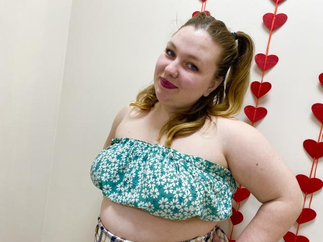 Una foto di Ailia con un corpo formoso e di carnagione chiara, che indossa un top a tubo floreale color turchese e bianco, sorridendo leggermente con le labbra rosse. Cuori sullo sfondo. I suoi capelli castani sono legati dietro.
.