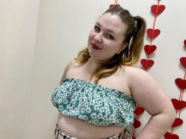 Ailia, een mollige vrouw met lichte huid en rood haar in staartjes, draagt een groene top met bloemen zonder bandjes. Rode, hartvormige decoraties hangen op de achtergrond. Ze kijkt naar de camera met een lichte glimlach..