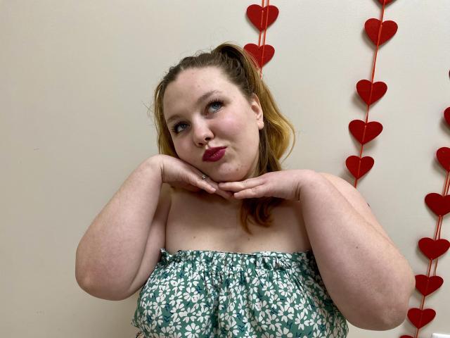 A photograph of Ailia with pale skin and brown hair in a ponytail, wearing a strapless green and white floral dress. She rests her chin on her hands, red heart decorations in the background. Fucking cute, playful expression.