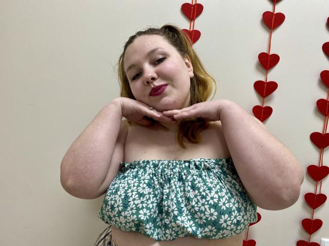 Ailia, a plus-sized woman with fair skin and light brown hair, wears a green floral strapless top, hands resting on her chin, looking down with a slight smile. Red heart decorations in the background.