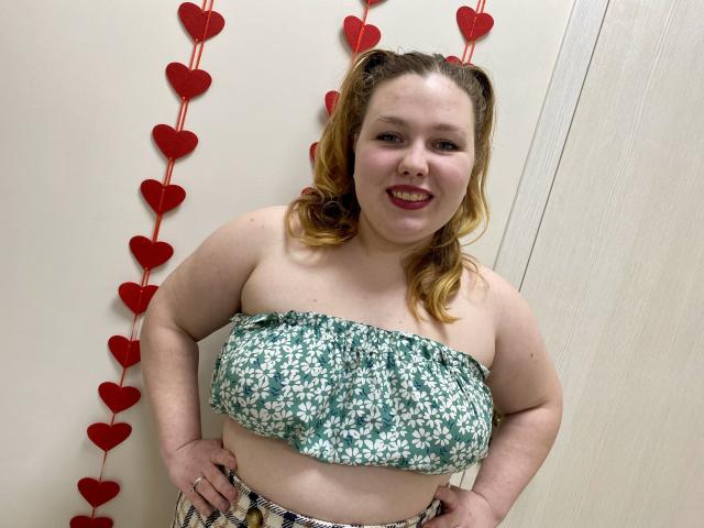 Ailia, una donna formosa con la pelle chiara e i capelli rossi, è in piedi, sorridente, con le mani sui fianchi, e indossa un abito floreale verde. top senza spalline, davanti a un muro bianco con decorazioni a forma di cuore rossi..