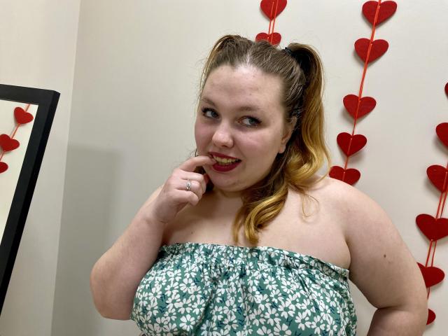 Ein Foto von Ailia mit heller Haut und braunen Haaren, die zu einem Pferdeschwanz gebunden sind, die ein grünes, blumiges Kleid ohne Träger trägt und dabei etwas in den Mund nimmt. Finger, lächelnd, mit roten, herzförmigen Dekorationen im Hintergrund..