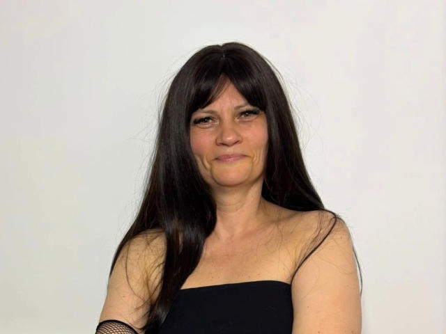 Photograph of LaraaFox-hot with long black hair, smiling slightly, wearing a black strapless top, white background, visible wrinkles on her face, slight sag in her breasts, gray border on top and bottom.
