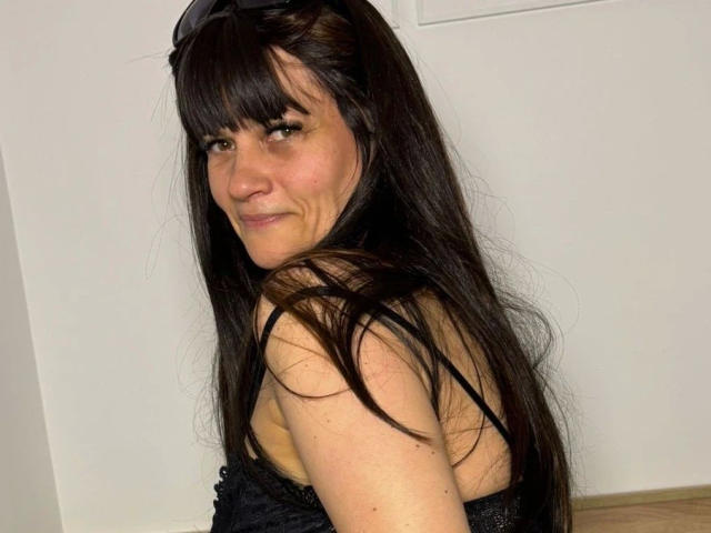 Photographie de LaraaFox, regardant légèrement de côté, cheveux noirs longs, frange, souriante, peau claire, haut noir sans bretelles, blanc fond mural, sol brun clair, tension sexuelle subtile, pose décontractée, lumière naturelle..