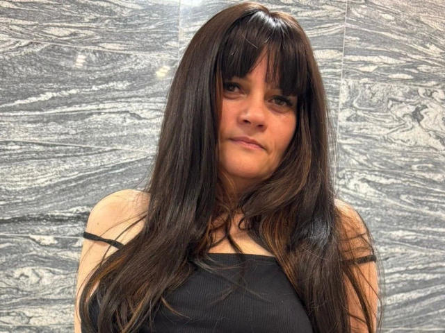 Fotografie von LaraaFox, attraktiv, mit langen, schwarzen Haaren, Pony und einem schwarzen Top mit Trägern, stehend vor einem strukturierten, silbergrauen Hintergrund. Hintergrund. Ihr Ausdruck ist leicht verführerisch, mit einem Hauch von sexueller Spannung..