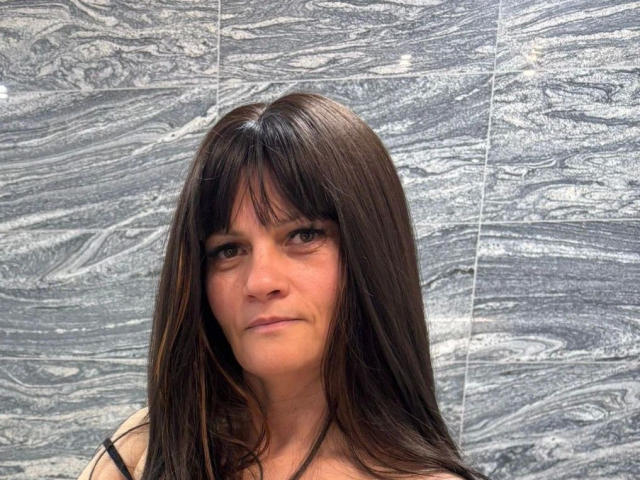 LarraaFox: mulher de meia-idade, com cabelos pretos longos, franja reta, expressão séria, parede de azulejos brancos com detalhes escuros. padrões ondulados em azul, leve sombra no lado direito, luz natural, tensão sexual sutil..
