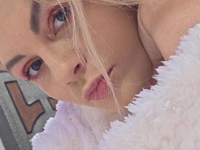 Close-up van Angelina-Joline, aantrekkelijk met roze oogschaduw, dikke eyeliner en rode lippen. Blond haar bedekt gedeeltelijk het gezicht, ze draagt een witte, pluizige badjas. Haar blik is intens en straalt seksuele spanning uit..