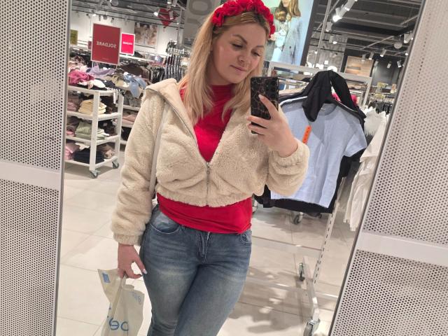 Fotografia di Angelina-Joline-hot che si scatta un selfie allo specchio in un negozio di abbigliamento. Indossa una maglietta rossa, una giacca bianca e pelosa, jeans blu e un foulard rosso, e tiene in mano una borsa della spesa. Sfondo: appendiabiti..