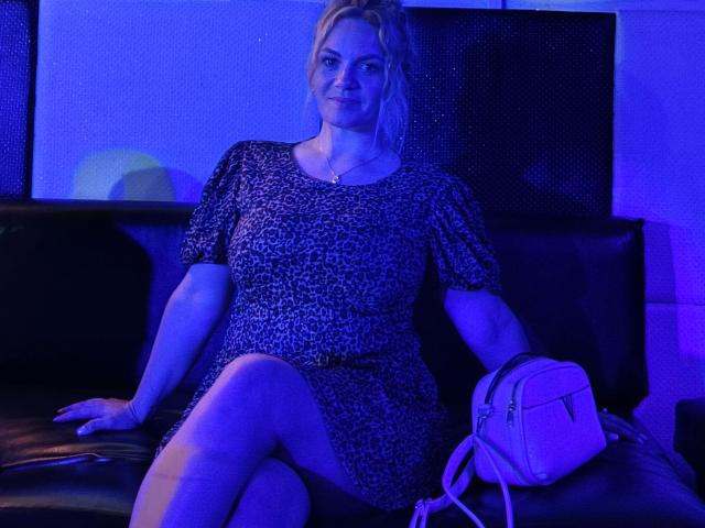 Foto con iluminación azul de Angelina-Joline-atractiva, sentada en un sofá negro, vistiendo una prenda de manga corta con un estampado blanco y negro. vestida, con las piernas cruzadas, mochila blanca a la derecha, mirando directamente a la cámara..