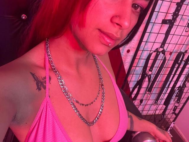 Primo piano di RoxanaSanchezz con illuminazione al neon rosa, che indossa una maglietta rosa scollata che mette in risalto il décolleté, e una catena d'argento. collana, tatuaggio a forma di farfalla sulla spalla sinistra. Sfondo: griglia nera e luci al neon. Tensione sessuale.