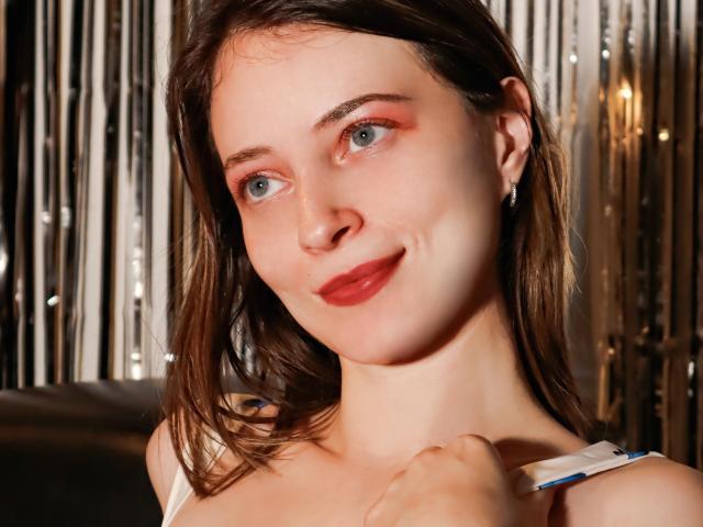 Nahaufnahme eines Fotos von AmbeerWood mit heller Haut, blauen Augen und rotem Lippenstift. AmbeerWoods braunes Haar reicht bis zu den Schultern, sie hält den Träger eines weißen Oberteils. Sonnenlicht betont das Gesicht und erzeugt Schatten..