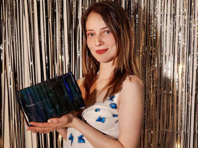 AmbeerWood está segurando uma clutch azul escura trançada, vestindo um vestido branco com estampas florais azuis. O fundo apresenta listras verticais brilhantes. Batom vermelho, cabelo castanho, sorriso confiante. Muito atraente..