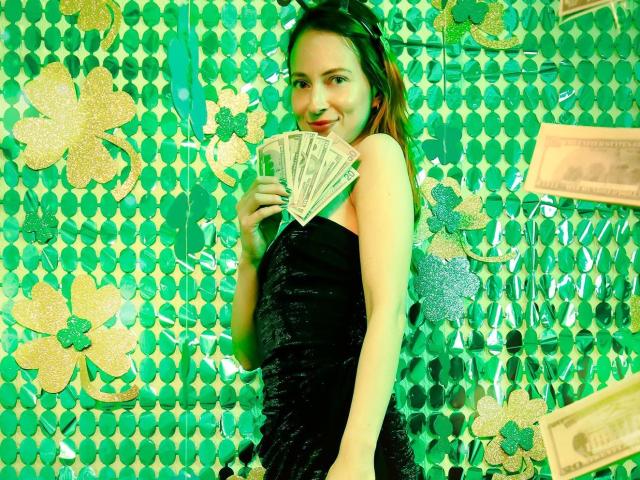 AmbeerWood, a young woman with fair skin and brown hair, stands against a green, patterned wall holding a fan. She wears a black dress, smiling with red lips, exuding sexual tension.