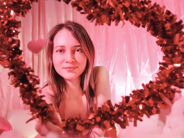 Photograph of AmbeerWood with wet brown hair, smiling, framed by red tinsel. Bright pink background. Red heart-shaped ornament on AmbeerWood's left. Sexual tension from direct gaze and intimate framing.