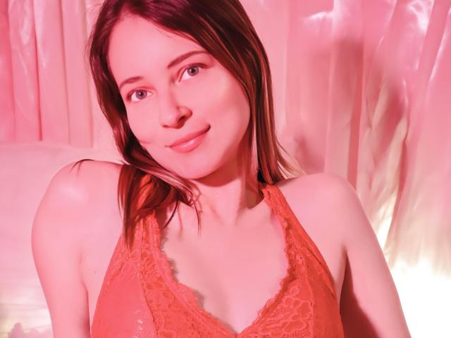 Fotografia ravvicinata di AmbeerWood con lunghi capelli castani, che indossa un reggiseno di pizzo rosso e sorride dolcemente. L'illuminazione rosa e bianca crea un'atmosfera calda e sensuale. Un'aria carnale ed erotica..