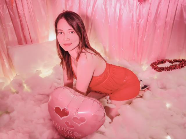 AmbeerWood, uma mulher de cabelos longos e escuros, ajoelha-se na neve fofa e branca, iluminada por uma luz rosa. Ela veste um vestido vermelho e segura um balão em formato de coração com a palavra "Amor" e símbolos de coração. Muito atraente..